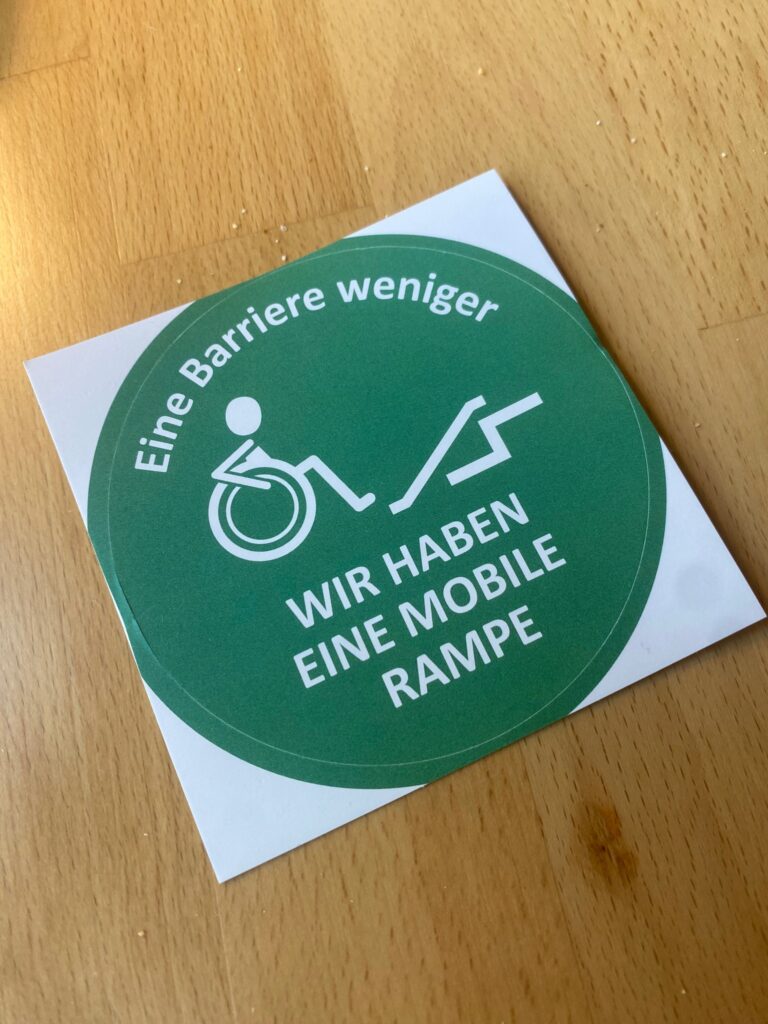 Mit der Rampe wird ein runder Aufkleber ausgegeben mit der Aufschrift "Eine Barriere weniger" - Wir haben eine mobile Rampe" und darauf ist eine Strichfigur zu sehen, die auf einer Rampe zwei Stufen hochfahren will.