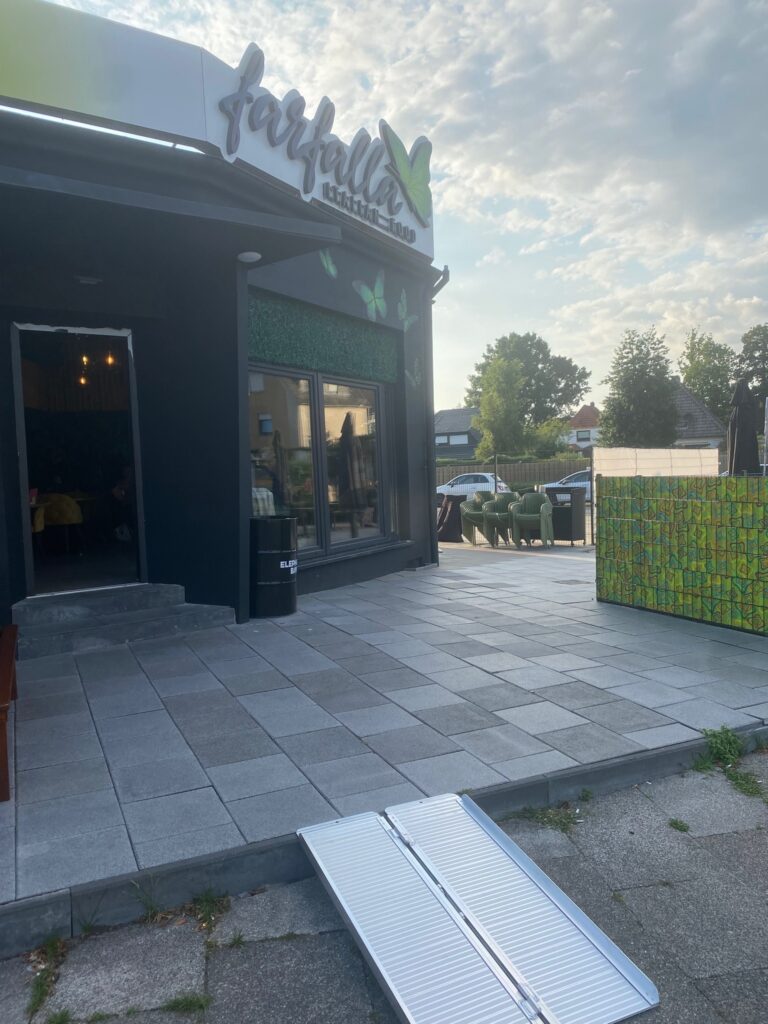 Wenn auf dem Bürgersteig für dem Lokal oder Geschäft genug Platz ist, muss die Rampe nicht immer bei Bedarf hingelegt und weggeräumt werden, sondern kann während der Öffnungszeit liegen bleiben, wie hier bei der ersten Stufe zum Lokal "Farfalla" in Huchting.