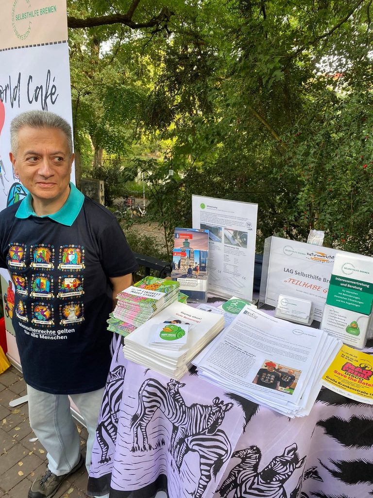Sepehr Dhegani mit dem T-Shirt mit 9 Forderungen der UN-BRK am LAGS-Stand (Stehtisch mit Zebra-Tischtuch und Material).